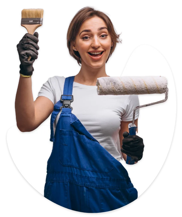 A cheerful female painter in blue overalls holding a paintbrush and a paint roller.
