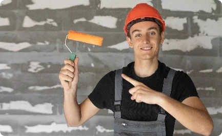 A young male painter in a red hard hat and overalls holding up and pointing to a paint roller.