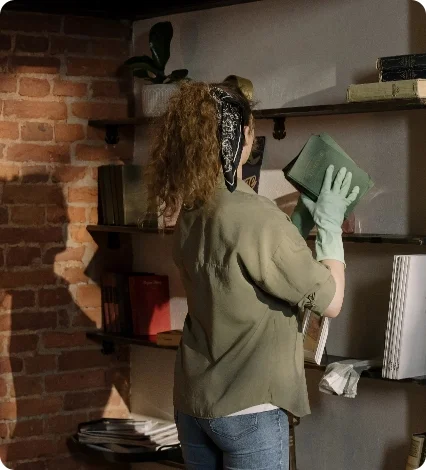 A woman wearing gloves carefully cleans and dusts books on a wooden shelf.