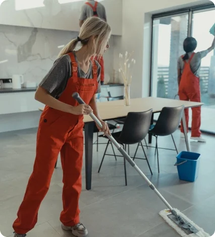 Two professional cleaners in matching orange uniforms mopping the floor and washing windows in a modern kitchen.