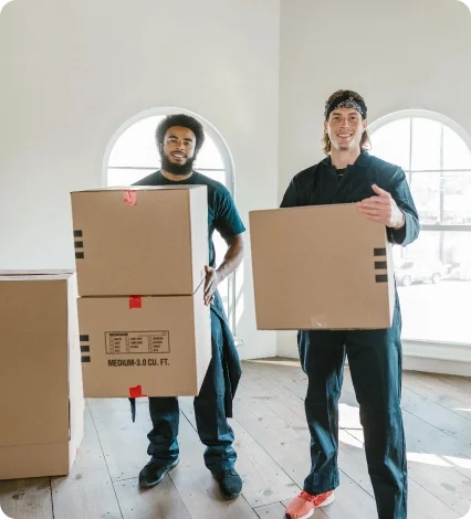 Two happy men standing on a wood floor in a bright, empty house with arched windows, holding moving boxes.
