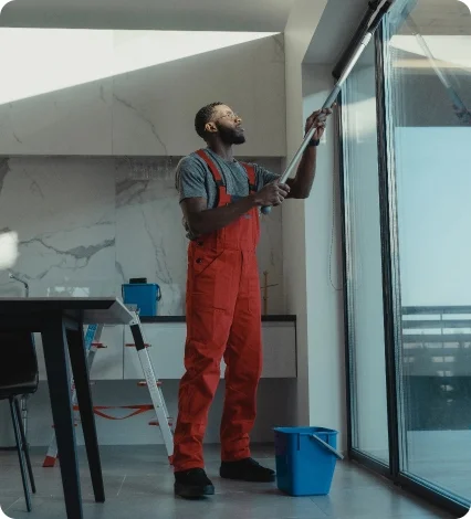 A man in orange overalls cleaning a large indoor window with a long-handled squeegee.