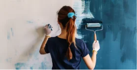 A woman seen from behind painting a wall with a roller of blue-grey paint.