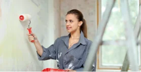 A woman smiling while painting an interior wall with a pink paint roller.